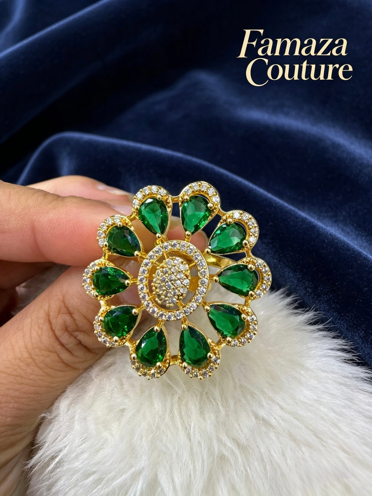 A gold-tone brooch finger ring featuring green teardrop-shaped gemstones and a central cluster of clear stones, held by a hand against a dark blue velvet background with a white faux fur surface. The brand name 'Famaza Couture' is visible in the top right corner.