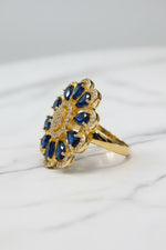 A gold-tone flower-shaped finger ring brooch with deep blue teardrop gemstones and a clear crystal centre, resting on a white textured faux fur surface.