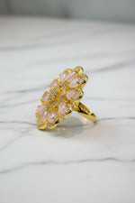 A gold-tone flower-shaped finger ring brooch with pale pink teardrop-shaped stones and a sparkling crystal centre, displayed on a white faux fur surface next to a brown textured bag.
