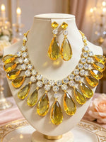 A gold-tone jewellery set featuring a bib-style necklace and matching drop earrings with large yellow teardrop gemstones and clear rhinestone accents, displayed on a cream jewellery bust against a softly blurred warm indoor background.