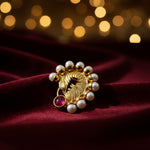 A gold-toned ornate brooch featuring a textured central motif, surrounded by small white faux pearls and accented with a round pinkish-red gemstone, shown on a plain light background.
