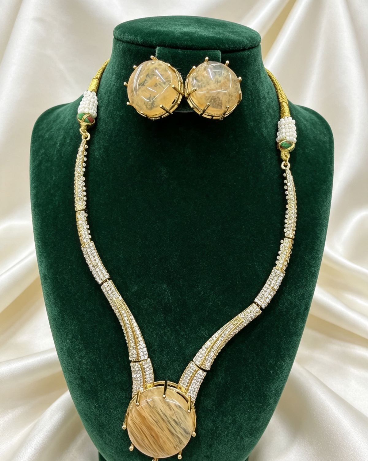 A gold and white beaded necklace set with a large oval pendant and matching stud earrings, displayed on a dark green velvet jewellery bust against a cream satin background.