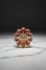 A gold flower-shaped brooch with pink teardrop gemstones and a central cluster of clear stones, held by a hand against a white furry background.