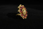 A gold flower-shaped finger Ring with pink teardrop gemstones and a textured gold centre, displayed on a white faux fur surface.