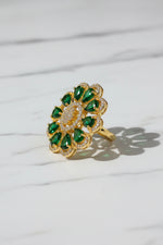 A gold flower-shaped finger ring brooch with green teardrop gemstones and a centre of small clear stones, held by a hand against a white faux fur background.