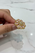 A gold flower-shaped finger ring brooch with pale pink teardrop stones and a central cluster of clear stones, held by a hand over a white textured surface.