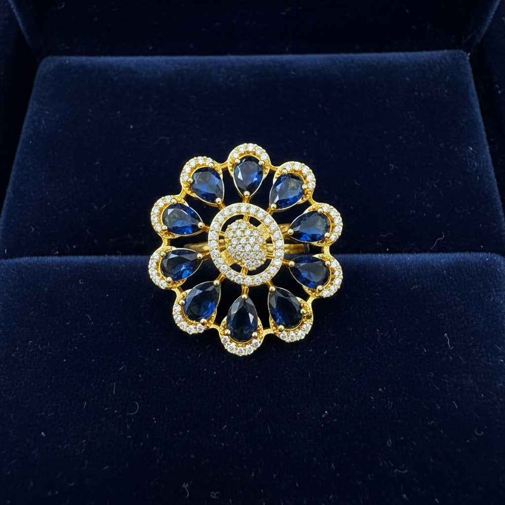 A gold flower-shaped finger ring with deep blue teardrop gemstones and a round centre paved with small clear stones, held between fingers against a white furry background.