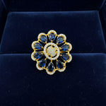 A gold flower-shaped finger ring with deep blue teardrop gemstones and a round centre paved with small clear stones, held between fingers against a white furry background.