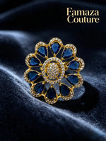 A gold flower-shaped ring with deep blue teardrop gemstones and a central cluster of small clear stones, displayed on a dark velvet surface. The brand name 'Famaza Couture' is visible in the top right corner.