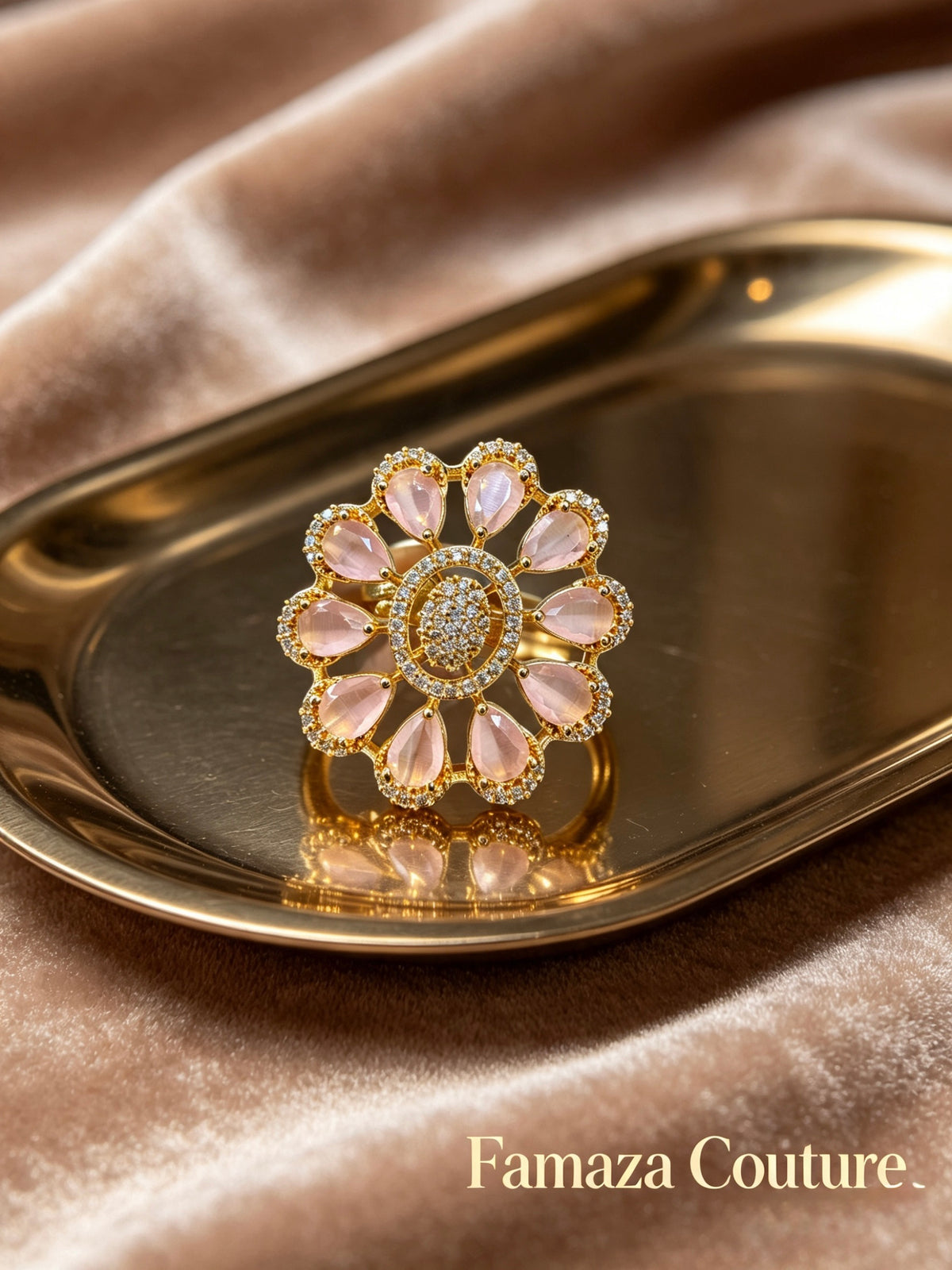 A gold flower-shaped ring with pale pink teardrop gemstones and a central clear stone, displayed on a metallic gold tray over a champagne-colored velvet fabric background. The brand name 'Famaza Couture' is visible in the bottom right corner.