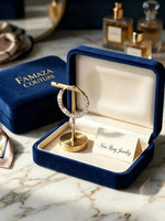 A gold ring stand holding a ring with clear stones, displayed inside an open navy blue jewelry box with a cream interior. A branded navy blue box labeled FAMAZA COUTURE sits beside it on a marble surface, with perfume bottles blurred in the background.