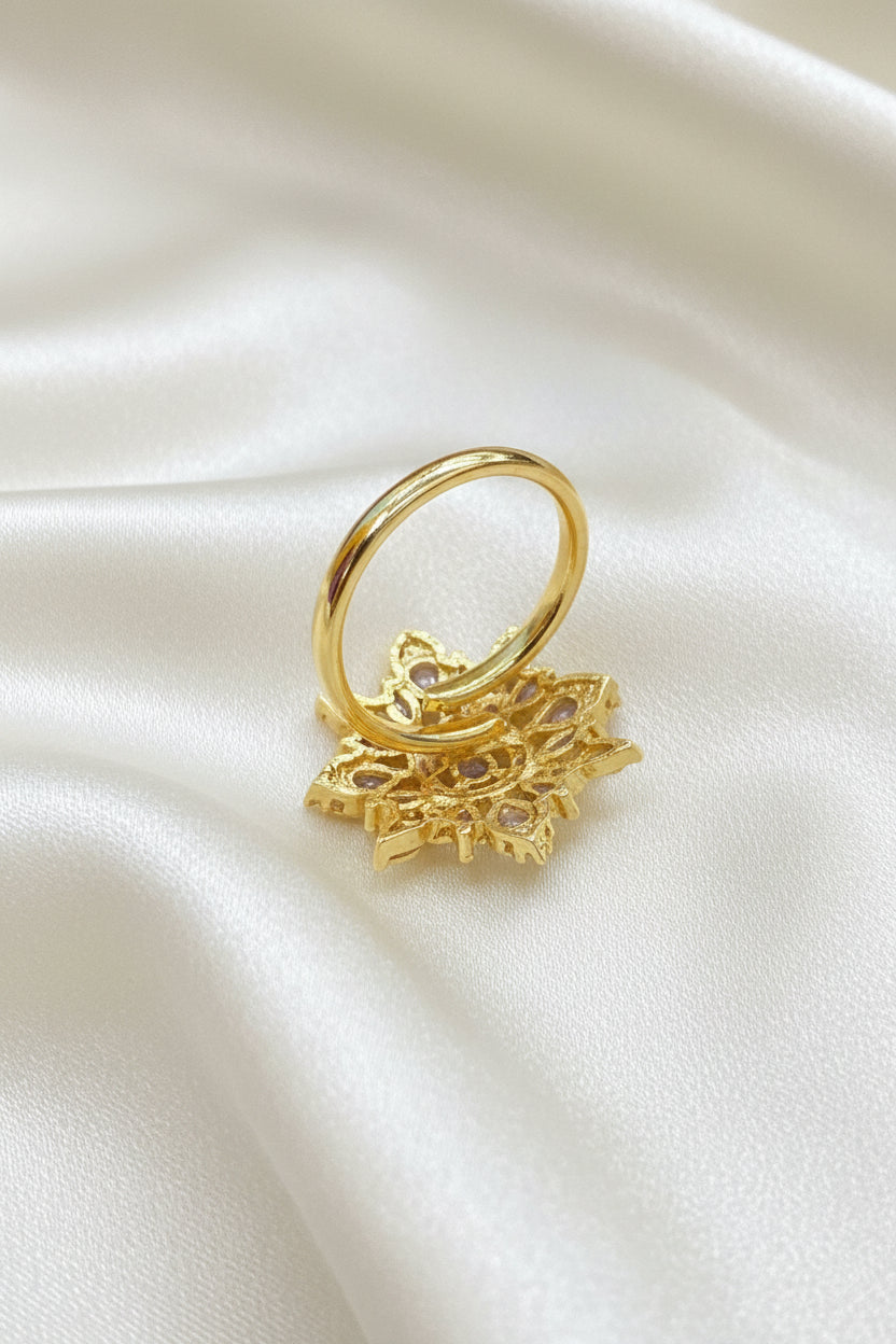 A gold ring with a large, intricate filigree snowflake or floral cluster setting, displayed on a textured blue velvet background.