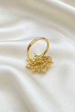 A gold ring with a large, intricate filigree snowflake or floral cluster setting, displayed on a textured blue velvet background.