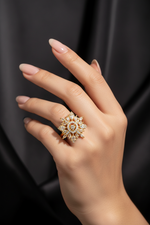A close-up of a hand wearing an ornate gold ring with clear gemstones, set against a dark, draped fabric background.