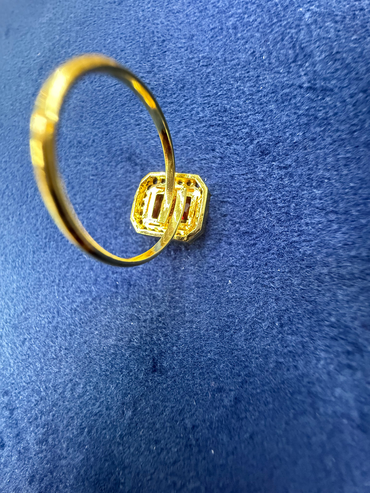 A gold ring with a textured, geometric square-shaped setting, displayed on a textured blue velvet surface.