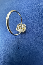 A silver-toned ring with a geometric, multi-layered square-shaped setting, photographed on a textured blue background.