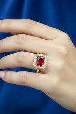 A hand holding a gold ring with a rectangular red gemstone surrounded by a halo of small clear stones, set against a blue velvet surface.