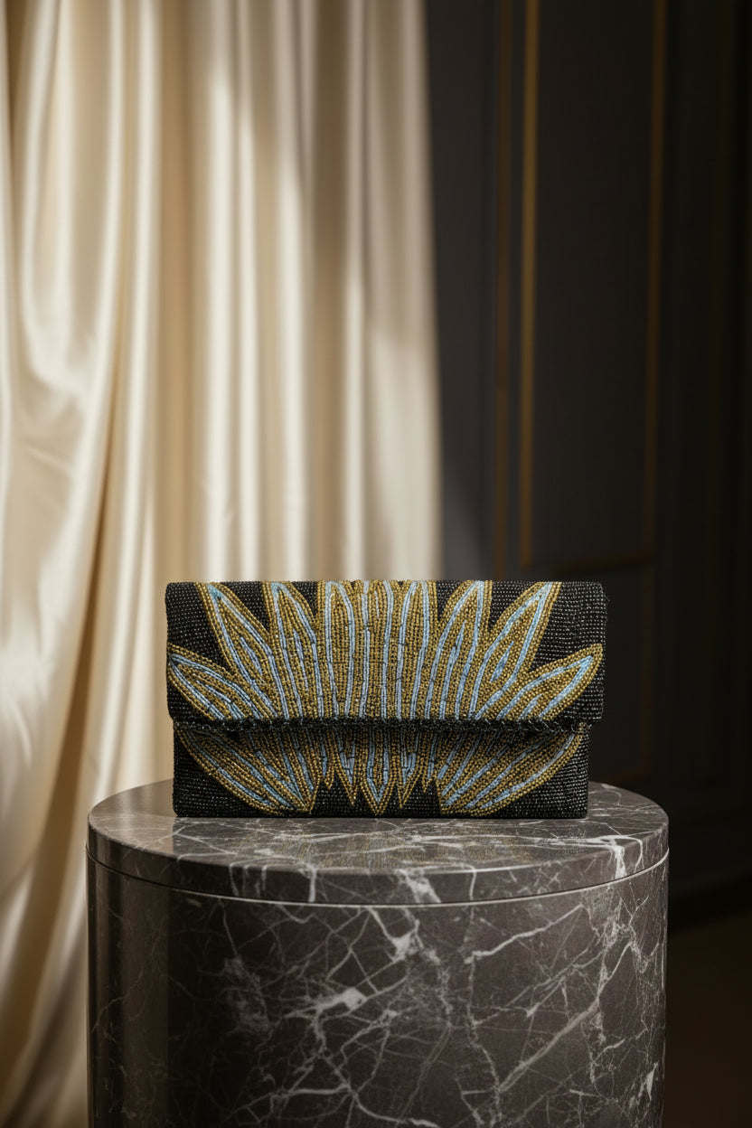 A handcrafted clutch bag with a geometric pattern of glass beads in gold and blue on a black base, displayed on a wooden surface with green leaf accents around.