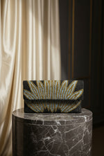A handcrafted clutch bag with a geometric pattern of glass beads in gold and blue on a black base, displayed on a wooden surface with green leaf accents around.