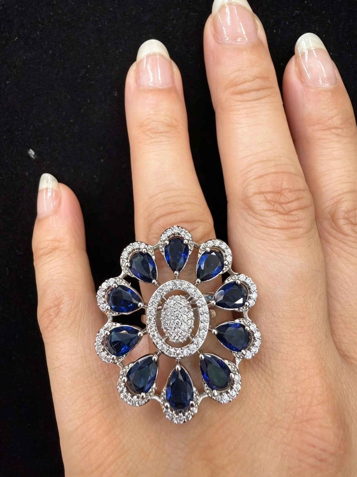A large floral cluster ring with deep blue teardrop-shaped stones and a silver-toned pavé center, shown worn on a hand against a black background.