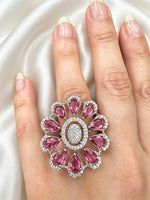 A large floral statement ring with pink teardrop gemstones and a central oval cluster of clear stones, shown worn on a hand against a satin fabric background.