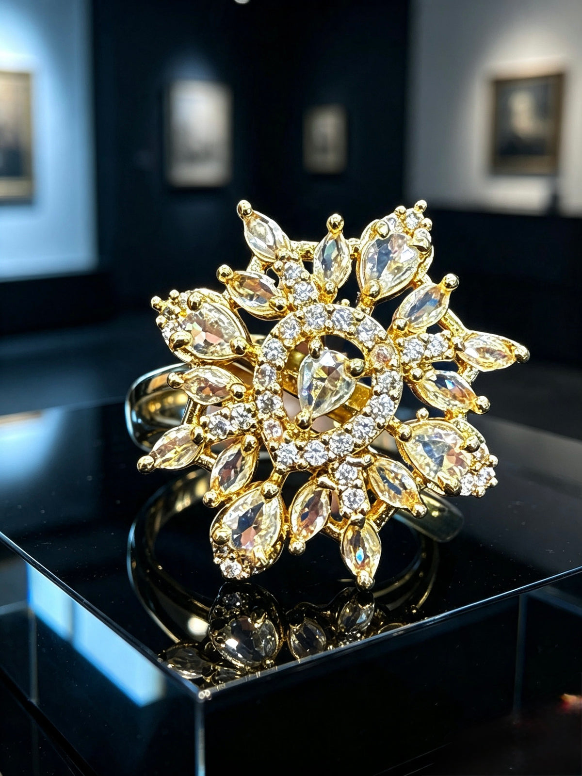 A large gold flower-shaped ring with marquise and round clear crystals, displayed on a glossy black surface in a dark showroom.