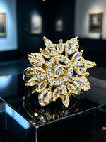 A large gold flower-shaped ring with marquise and round clear crystals, displayed on a glossy black surface in a dark showroom.
