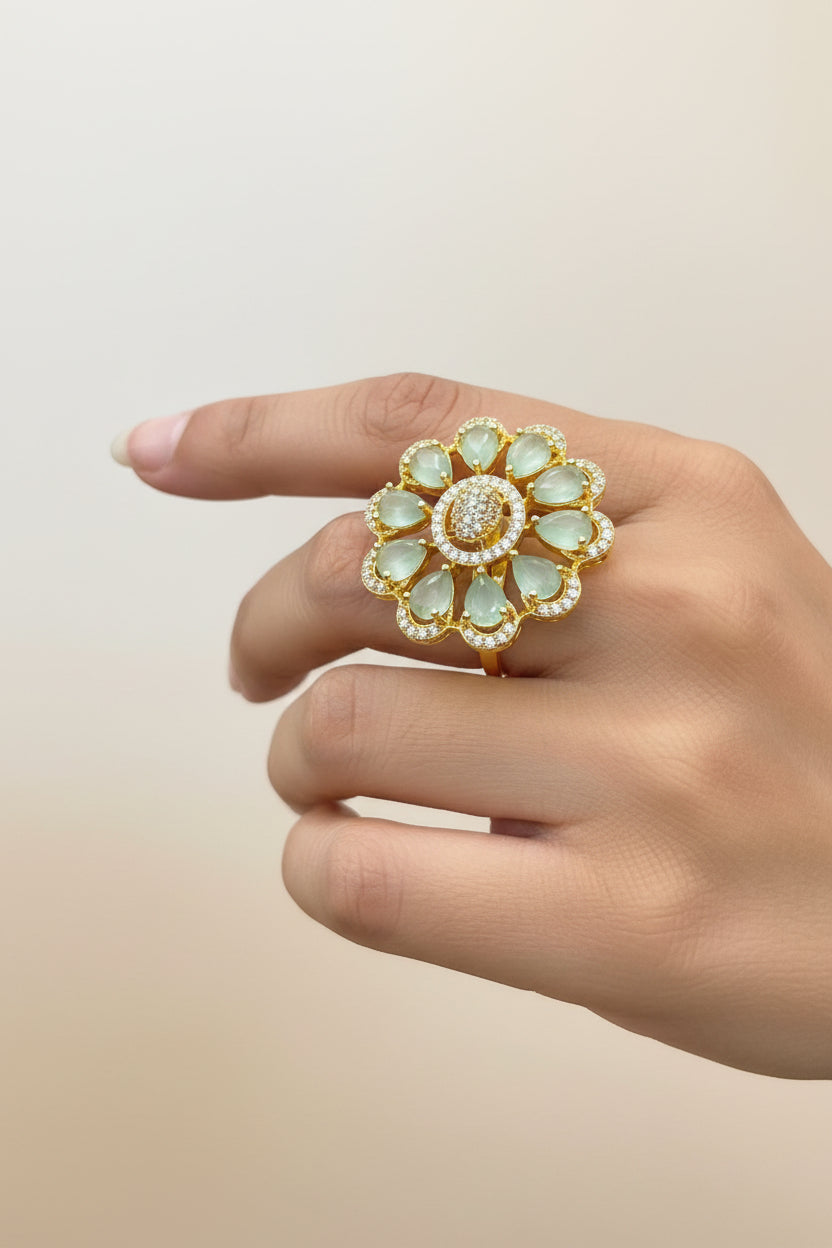 A large gold flower-shaped ring with pale green teardrop gemstones and a central cluster of small clear stones, shown worn on a person's finger.