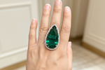 A close-up of a hand wearing a large teardrop-shaped emerald-cut green gemstone ring with a silver-toned halo set with small clear stones, against a softly blurred indoor background.