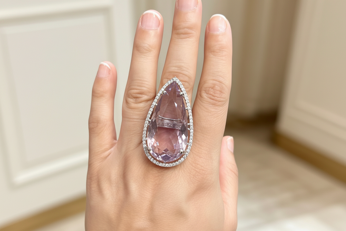 A close-up of a hand wearing a large teardrop-shaped lavender-purple gemstone ring with a halo of small clear stones, set against a softly blurred indoor background.