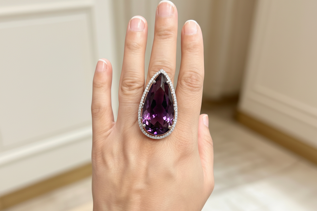 A close-up of a hand wearing a large teardrop-shaped purple gemstone ring with a silver-toned halo and small accent stones, set against a softly blurred indoor background.