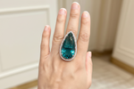 A close-up of a hand wearing a large teardrop-shaped emerald-cut blue gemstone ring with a halo of small clear stones, set against a softly blurred indoor background.