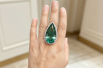A close-up of a hand wearing a large teardrop-shaped green gemstone ring with a silver-toned halo and small clear accent stones, set against a softly blurred indoor background.
