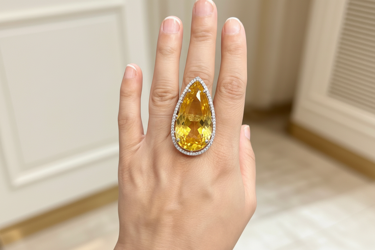 A close-up of a hand wearing a large teardrop-shaped yellow gemstone ring with a halo of small clear stones, set against a softly blurred indoor background.
