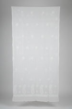 A large white sheer fabric panel or curtain with subtle tonal floral embroidery, hanging in a room with a window and mannequin visible in the background.