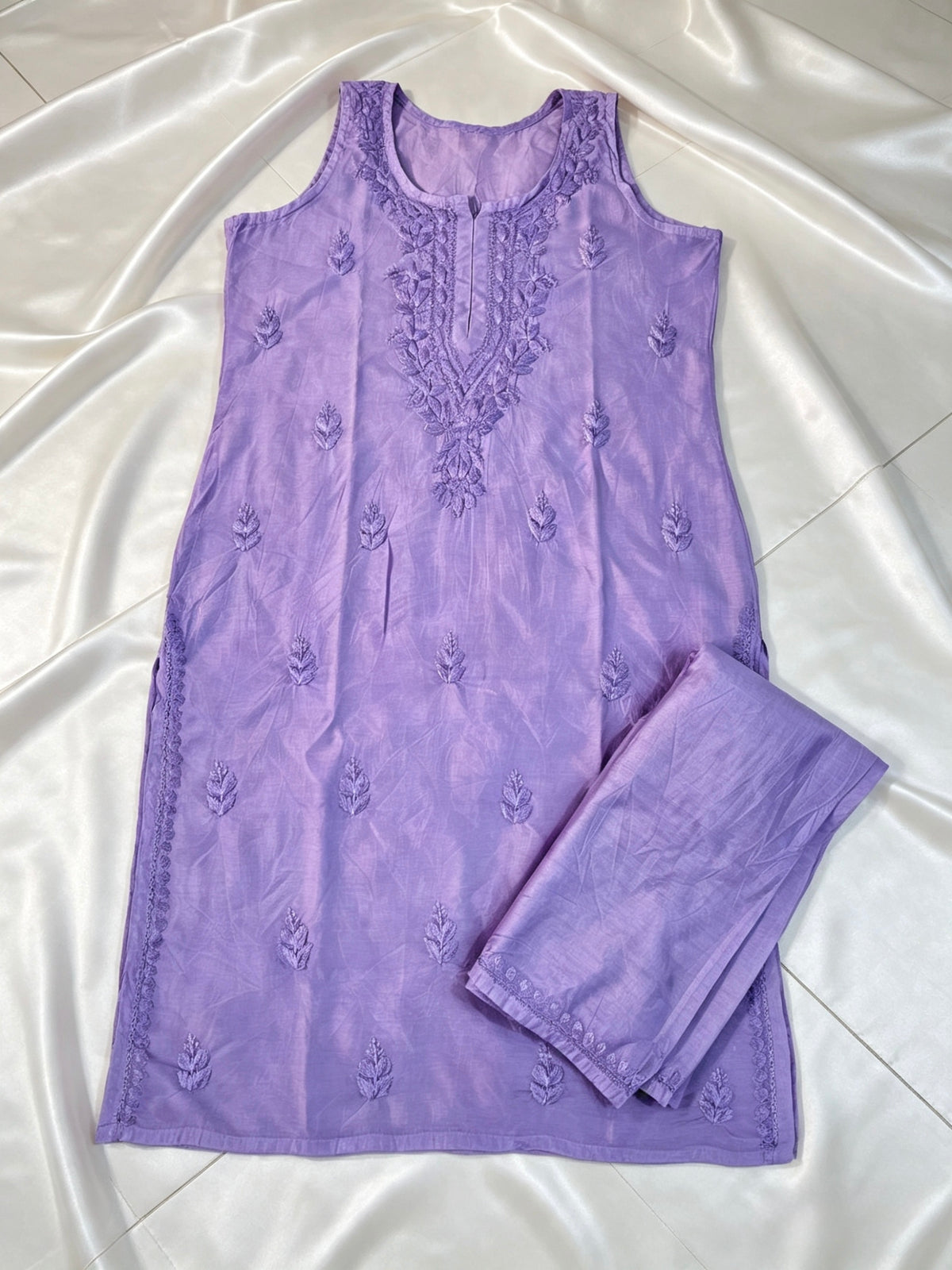 A lavender purple sleeveless kurta with floral embroidery on the neckline and scattered embroidered motifs across the front, paired with matching folded pants, laid flat on a white satin fabric background.