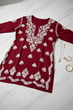A long-sleeve maroon tunic top with white floral and leaf embroidery, hanging on a wooden hanger against a plain white background.