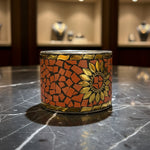A mosaic cuff bracelet featuring a sunflower design with orange and gold tile patterns, photographed on a dark textured surface.