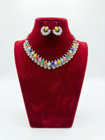 A multi-color gemstone necklace featuring orange, blue, green, and pink stones in silver-plated filigree, shown with yellow stud earrings on a red bust.