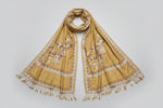 A mustard Kashmiri Aari embroidered marina scarf with tassels hanging at the ends, displayed on a wooden hanger with greenery in the background.