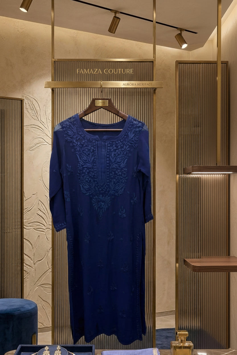 A navy blue embroidered dress displayed on a hanger inside a luxury boutique, with jewellery and a folded matching blue scarf on a marble counter below.