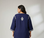 A navy blue long-sleeve tunic or kurta with white embroidered details on the chest and sleeve cuffs, laid flat on a gray herringbone wood floor.