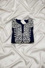 A navy blue tunic top with intricate white embroidery and floral motifs, laid flat on a grey wood floor with a white tag still attached at the collar.