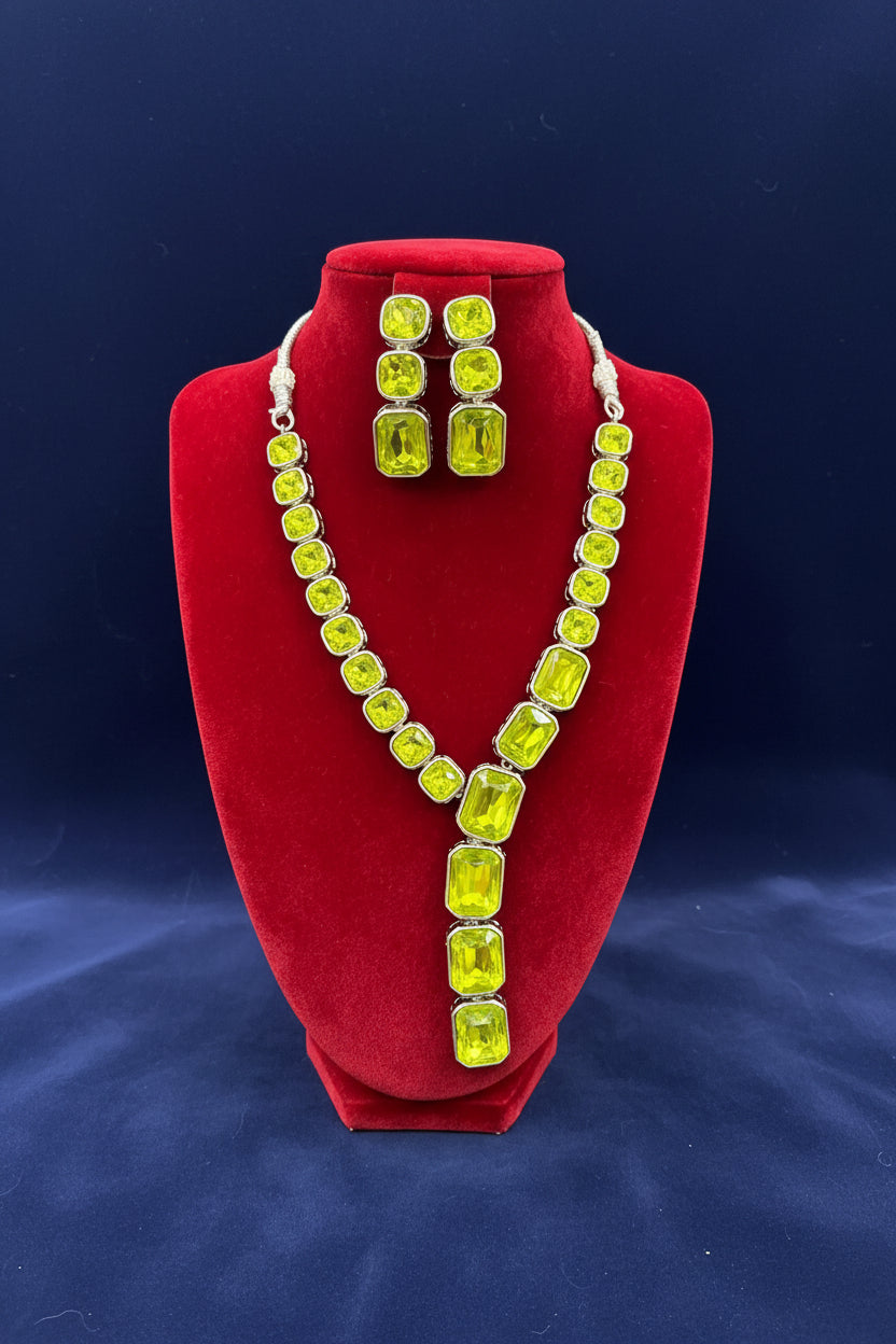 A neon green rhinestone necklace and matching drop earrings displayed on a red velvet jewellery bust, with a busy workshop background.