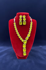 A neon green rhinestone necklace and matching drop earrings displayed on a red velvet jewellery bust, with a busy workshop background.
