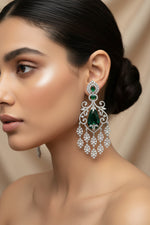 A pair of American diamond ornate silver-toned chandelier earrings with large green teardrop-shaped center stones and smaller green stones above, embellished with clear rhinestones, displayed on a light wood surface.