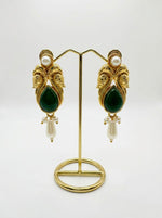 A pair of deep bottle-green jhumka earrings displayed on a stand, highlighting the rich, polished green cabochon center against an intricate, vintage-inspired brass setting with hanging pearl accents.