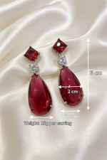 A pair of long drop earrings with deep red teardrop-shaped gemstones, square red stones at the top, and clear round stones in between, displayed on a cream satin background with measurement annotations showing 6 cm height, 2 cm width, and 15g weight per earring.