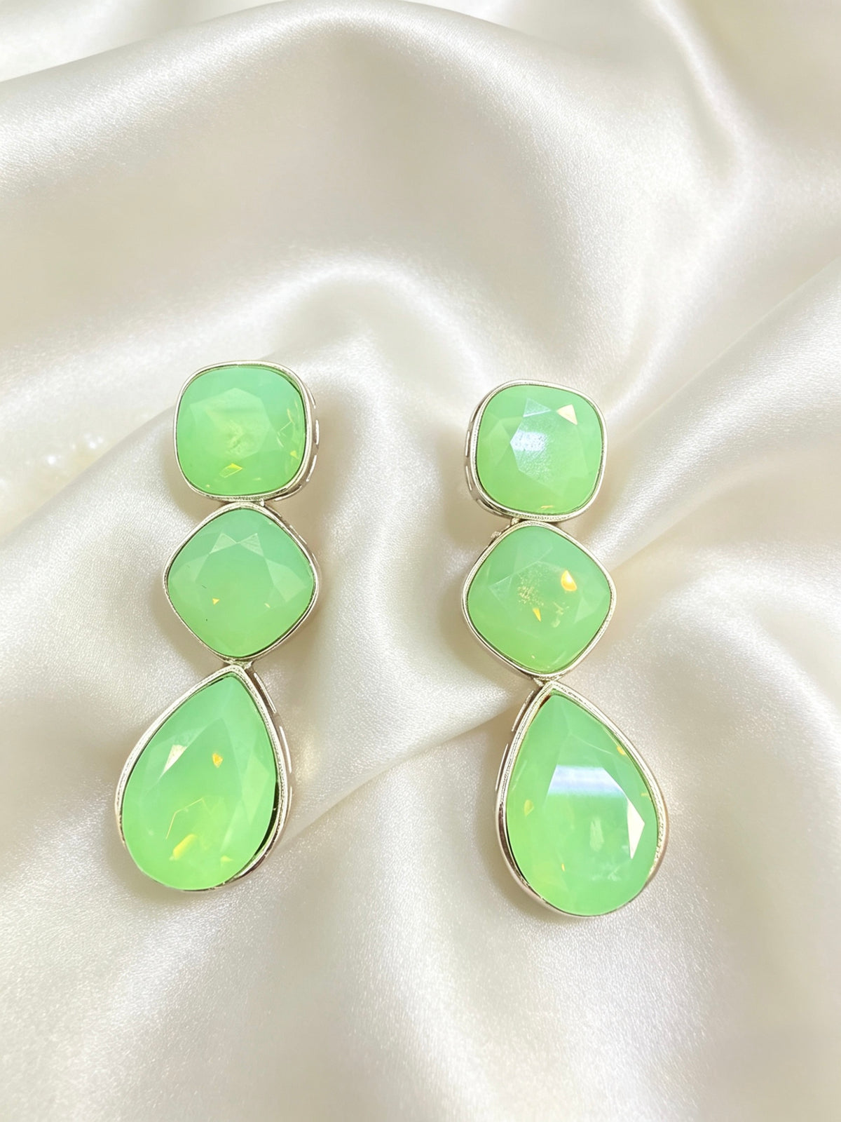 A pair of drop earrings with three stacked light green faceted stones in silver-toned settings, displayed on a cream satin fabric background.