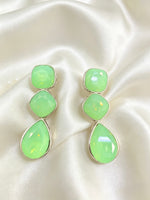A pair of drop earrings with three stacked light green faceted stones in silver-toned settings, displayed on a cream satin fabric background.